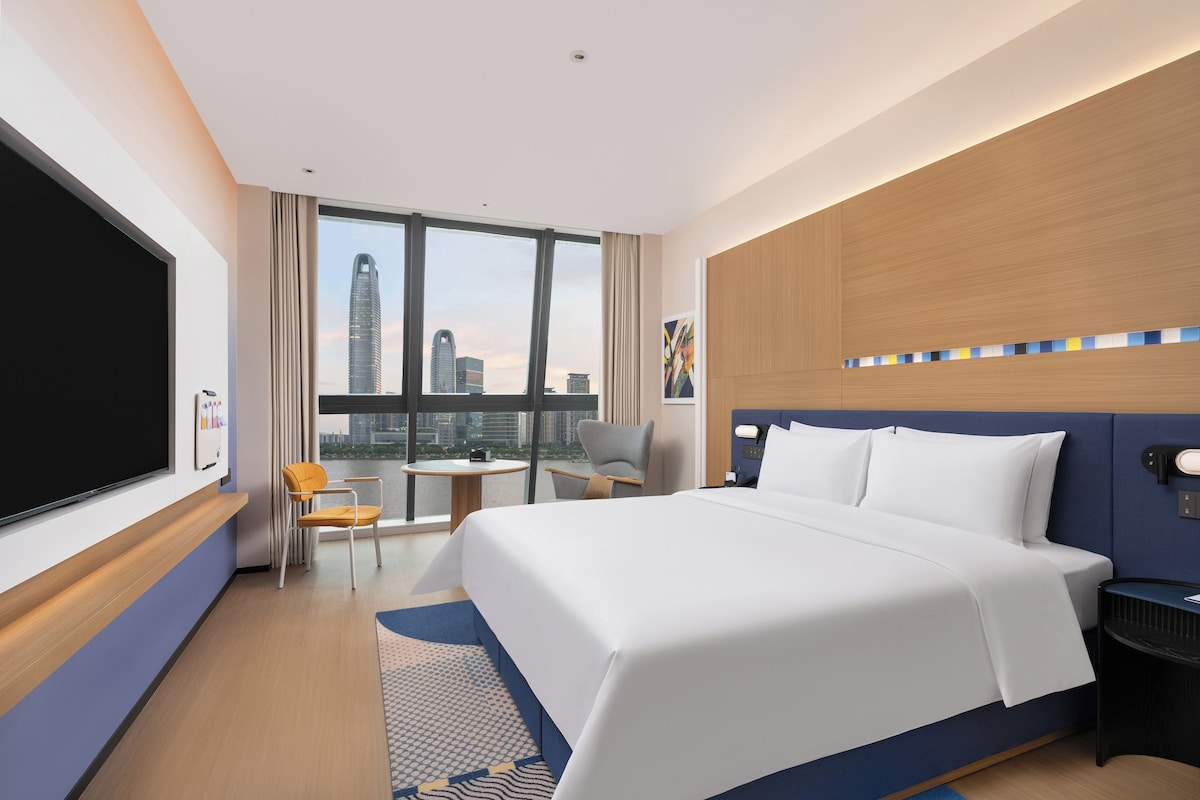 A hotel room with a large bed, a desk, a chair, and a view of the cityscape.