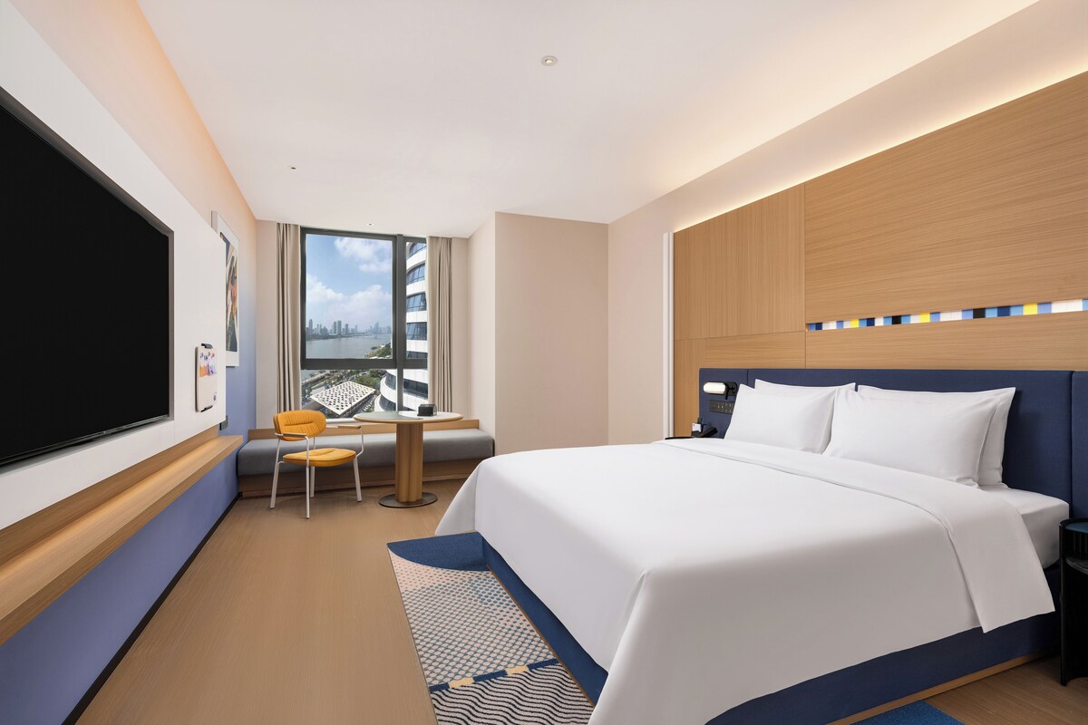 A hotel room with a large bed, a desk with a chair, a TV, and a window with a city view.