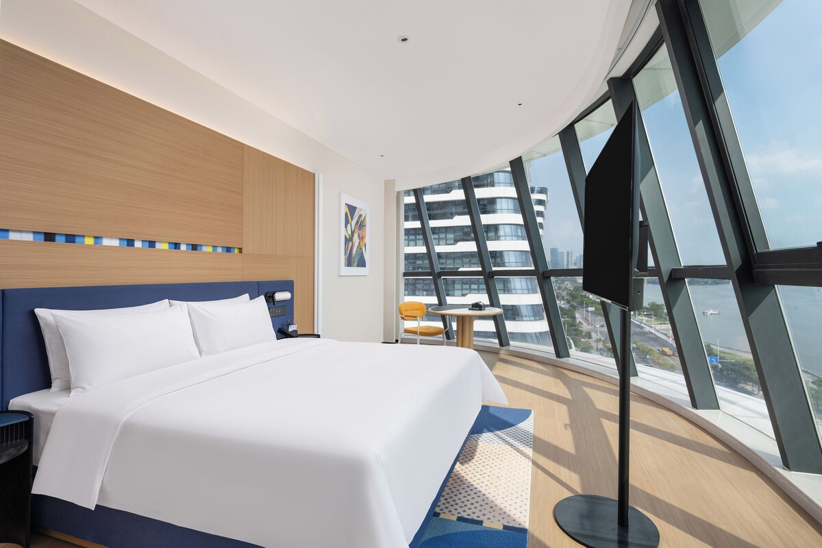 A modern hotel room with a large bed, a desk, a chair, and a view of the city through large windows.
