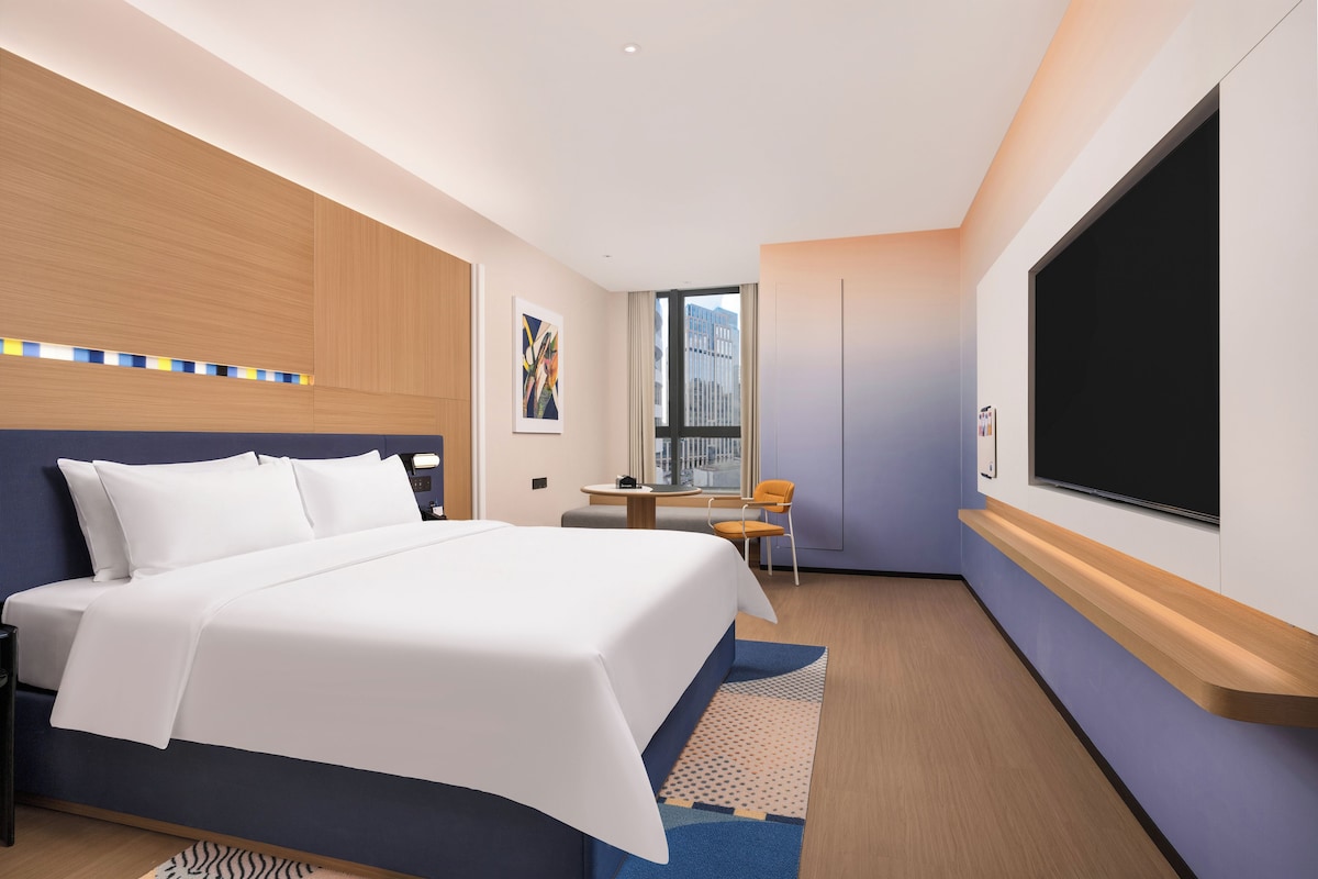 A modern hotel room with a large bed, a desk, a chair, and a TV.