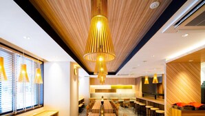 A modern dining area with wooden tables and chairs, a central hanging light fixture, and a kitchen in the background. - Super Hotel Tokyo Shiba (Tokyo)