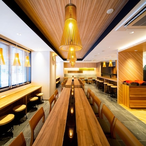 Un espace de restauration moderne avec des tables et des chaises en bois, un luminaire suspendu au centre et une cuisine en arrière-plan.