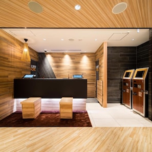 Un área de recepción con un mostrador de madera, una computadora y una caja registradora. Hay dos bancos de madera y una pared oscura con el letrero 'Super Hotel'.