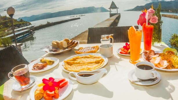 A table set with breakfast items, including waffles, eggs, and fruit, overlooking a body of water with a dock and mountains in the distance. - Toba Village Inn (Samosir)