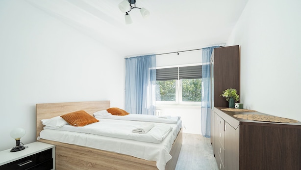 A bedroom with a bed, bedside table, lamp, and a view of the outdoors. - RentPlanet - Apartament Górczewska (Warszawa)