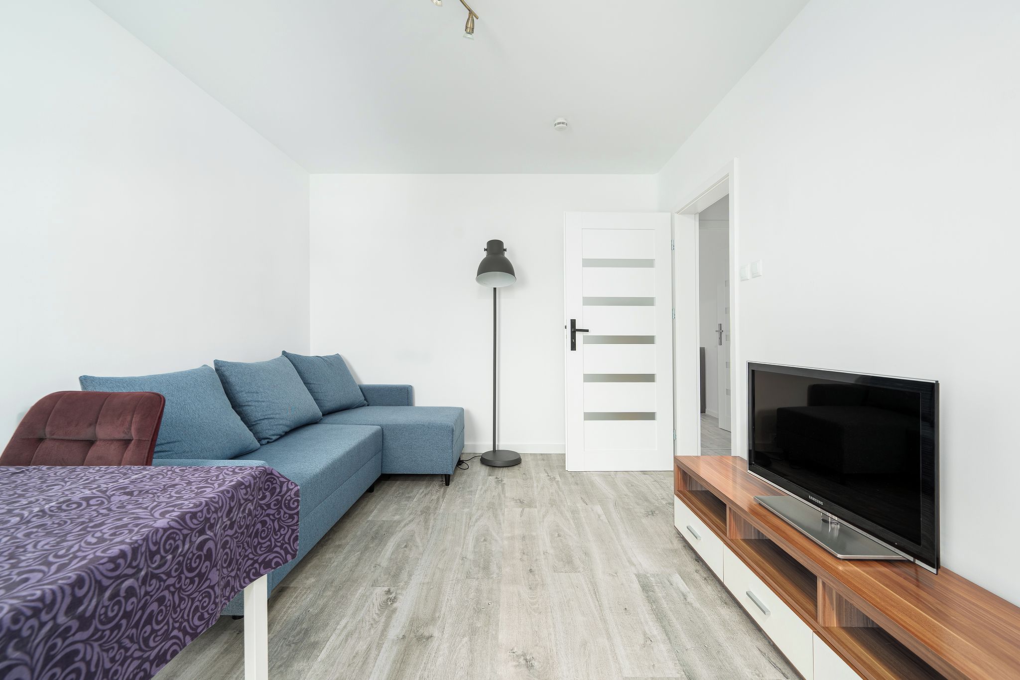 Een moderne, compacte leefruimte met een bank, een televisie op een houten dressoir, een vloerlamp en een deur naar een andere kamer.