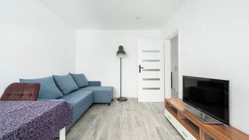 Een moderne, compacte leefruimte met een bank, een televisie op een houten dressoir, een vloerlamp en een deur naar een andere kamer.