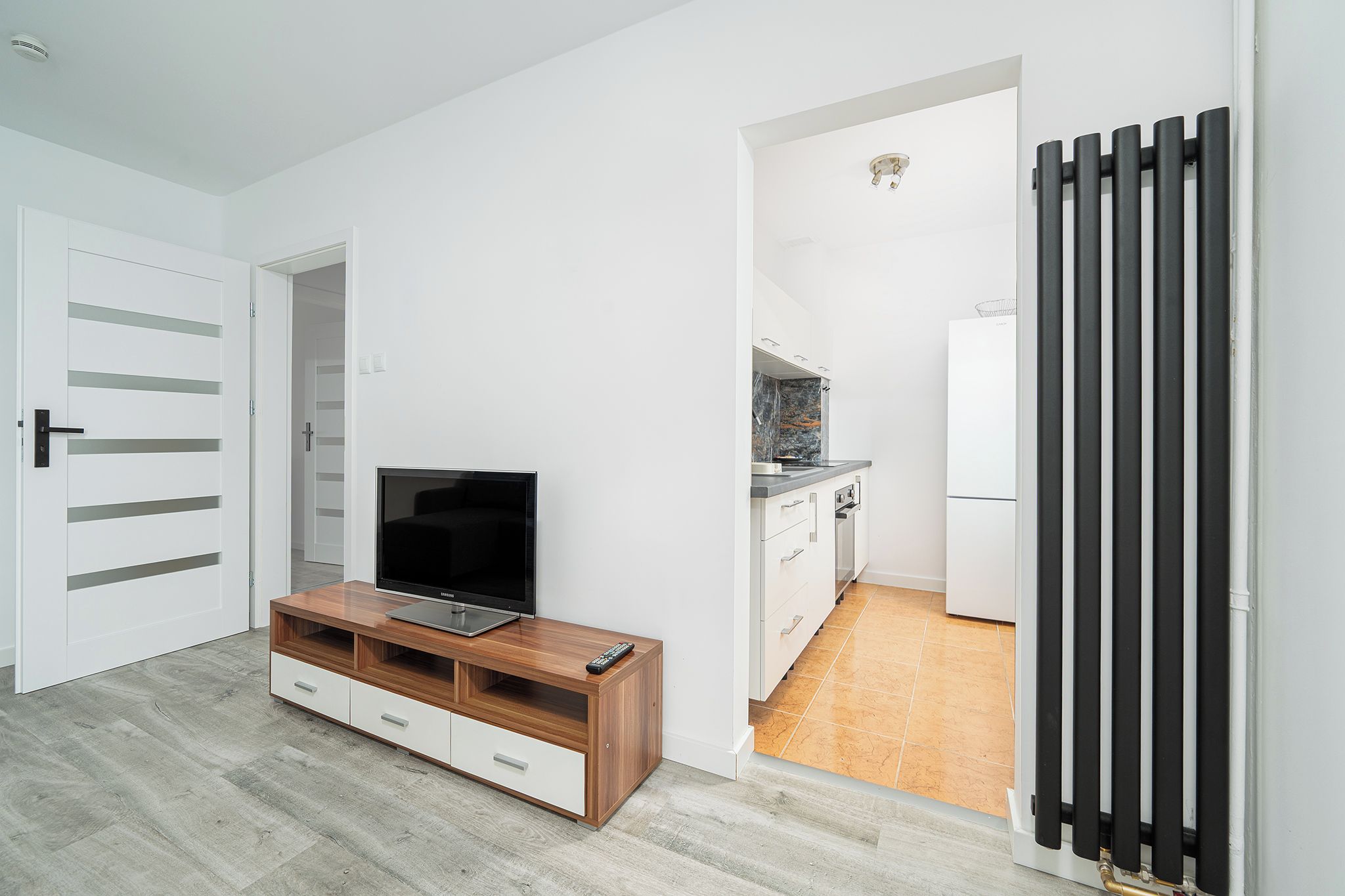 Een moderne woonkamer met een televisie op een houten dressoir, een witte deur en een zichtbare keuken door een open doorgang.