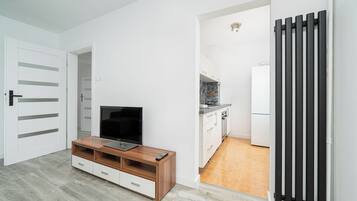Een moderne woonkamer met een televisie op een houten dressoir, een witte deur en een zichtbare keuken door een open doorgang.