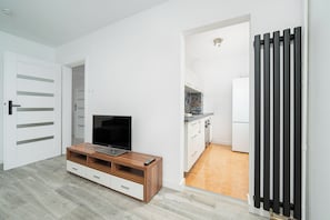 Un saló modern amb un televisor sobre un suport de fusta, una porta blanca i una cuina visible a través d'una obertura.