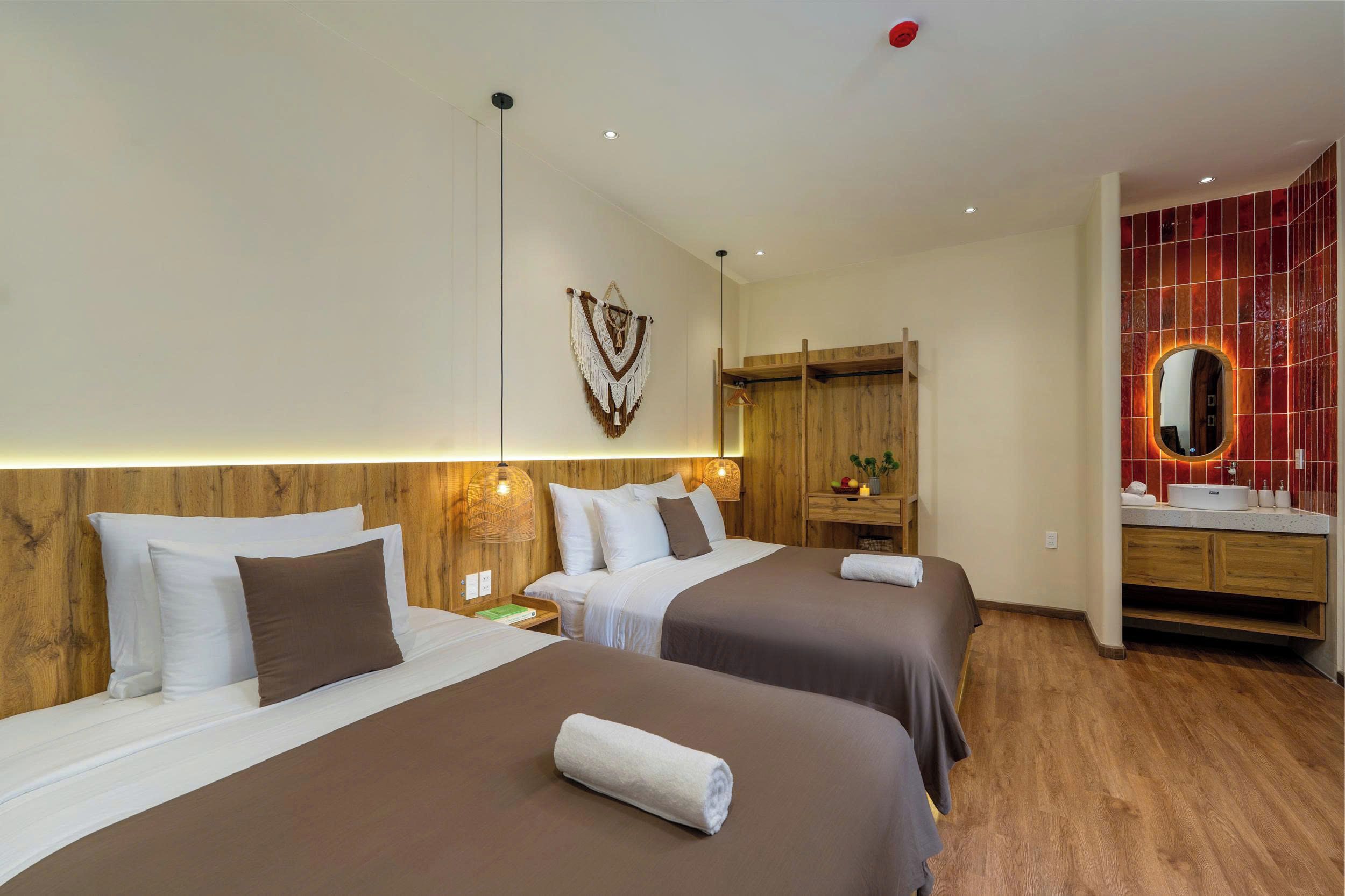 A hotel room with two beds, a wooden headboard, a wall-mounted macrame decoration, a wooden wardrobe, a vanity with a mirror, and a red-tiled wall.