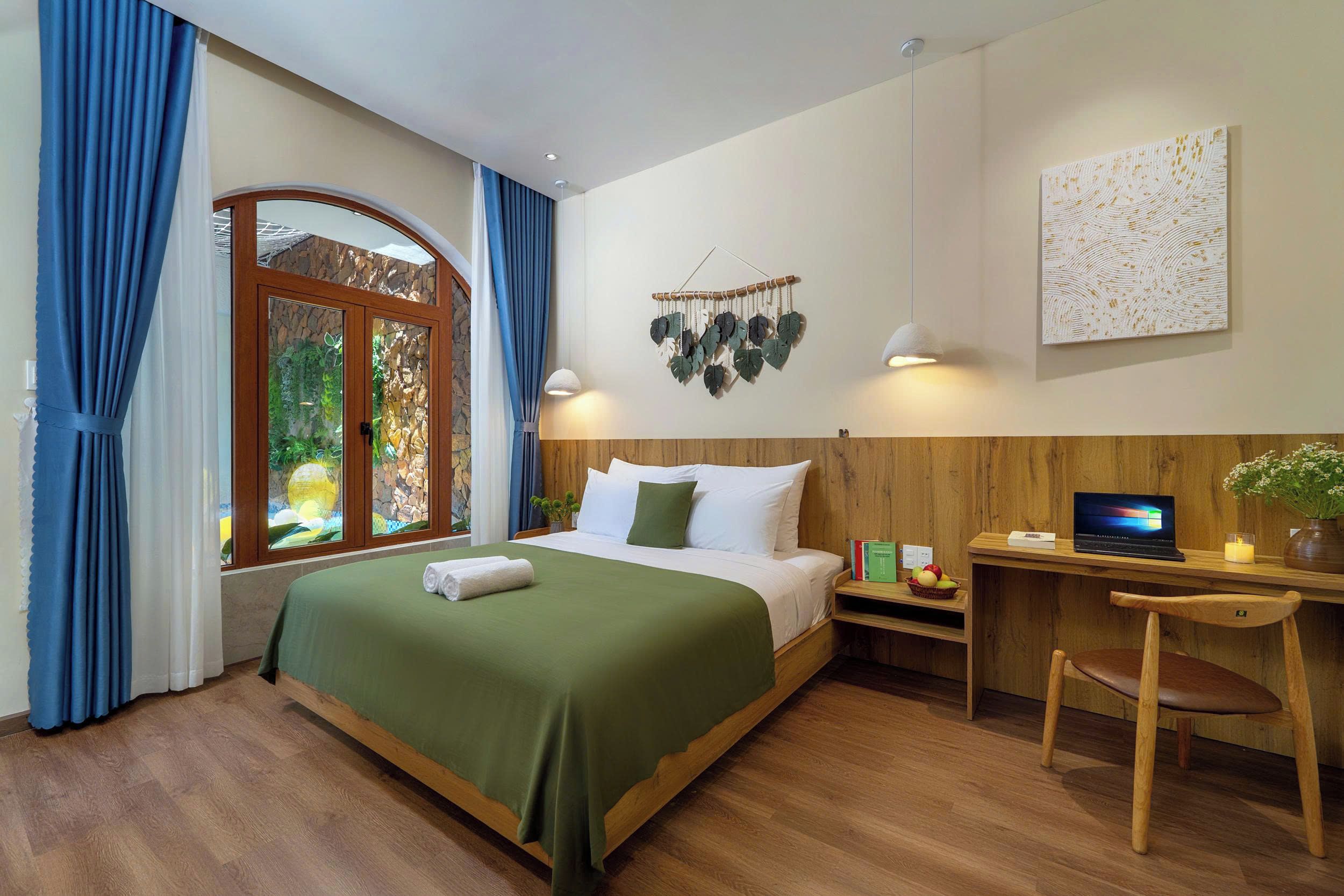 A hotel room with a bed, a desk with a laptop, a chair, a wall-mounted plant, and a window with curtains.