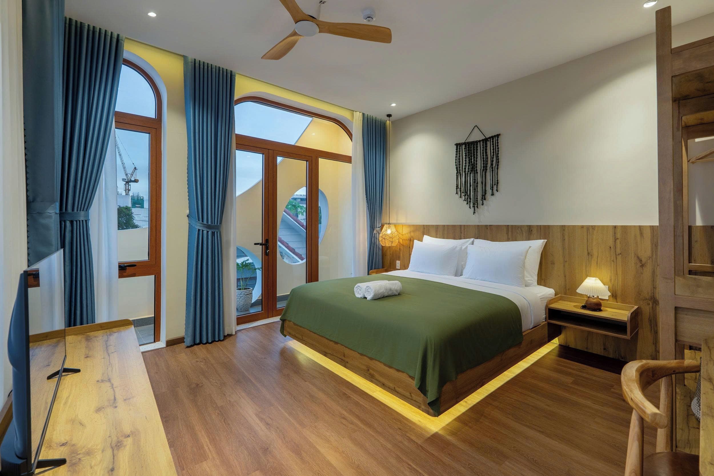 A hotel room with a bed, wooden flooring, a ceiling fan, a nightstand with a lamp, and a view of the outside through a glass door.