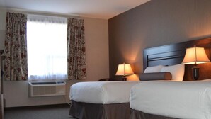 A hotel room with two beds, a window with curtains, a bedside table with a lamp, and a wall-mounted heater. - Cobble Creek Lodge (Maple Creek)