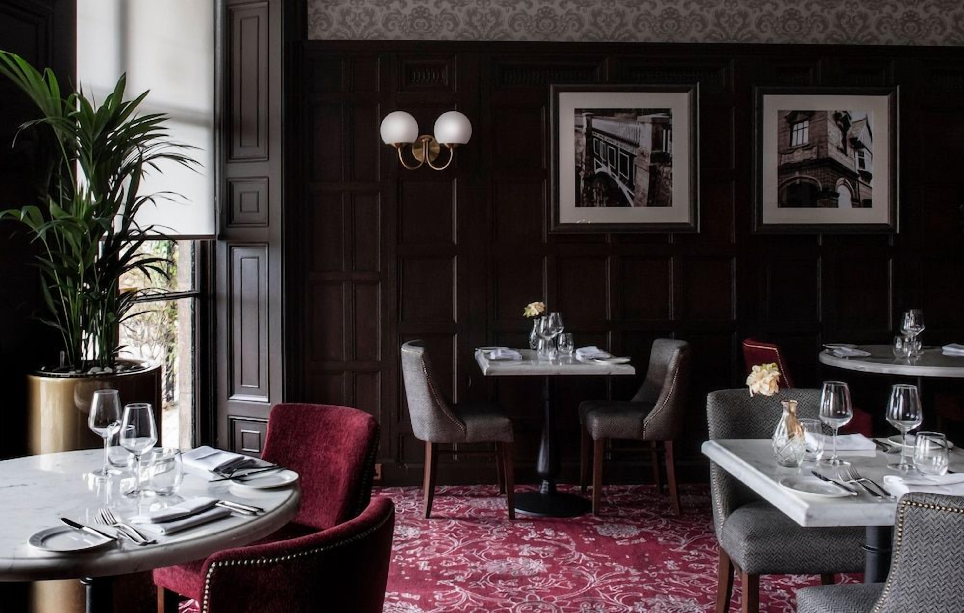 A dining area with tables set for two, a potted plant, and framed pictures on the wall.