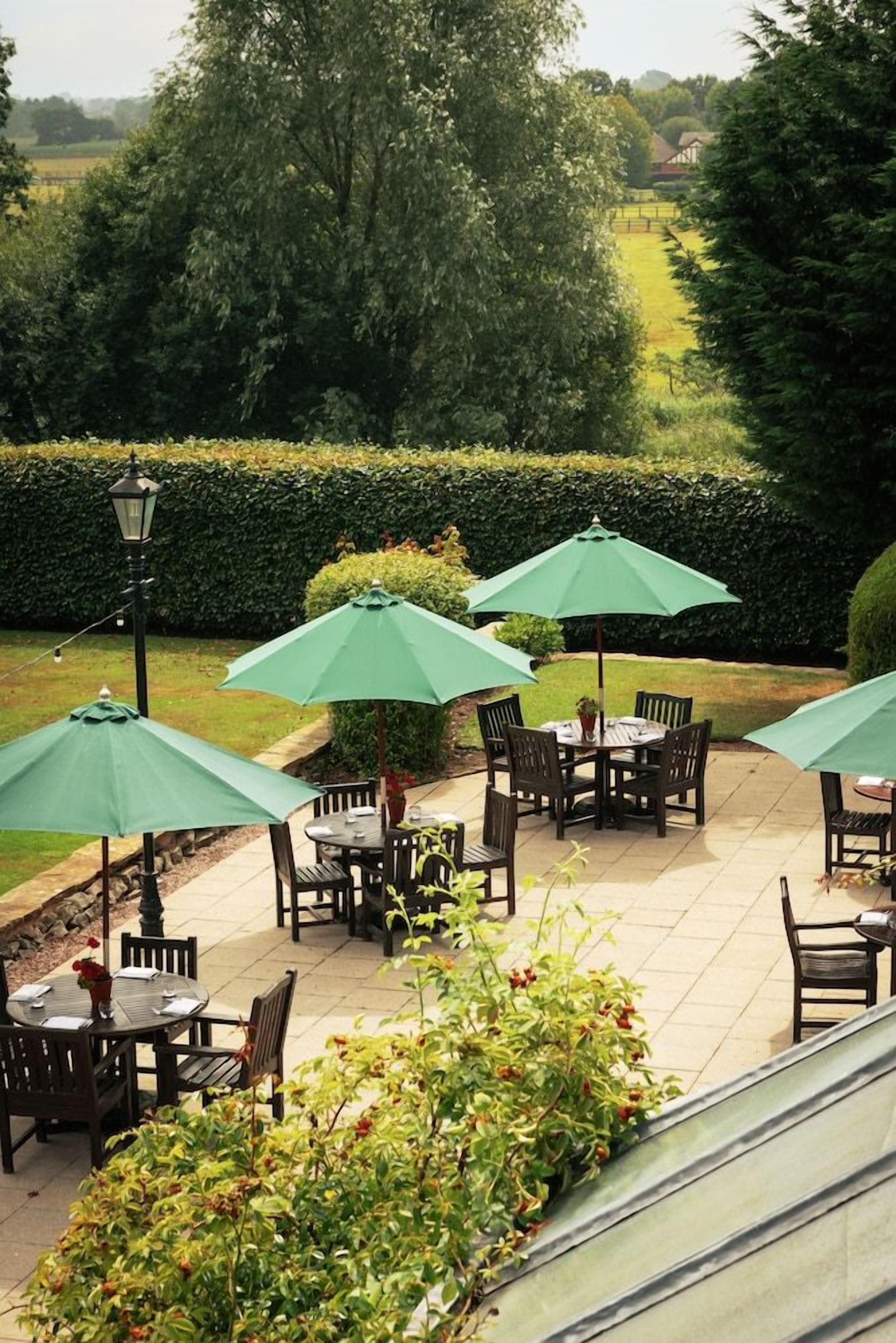 An outdoor patio area with tables and chairs, surrounded by greenery and a hedge.