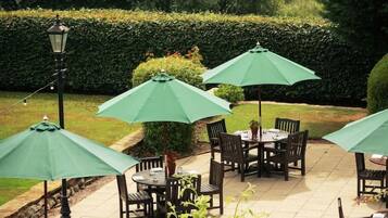 An outdoor patio area with tables and chairs, surrounded by greenery and a hedge.