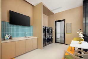A hotel room with a flat-screen TV mounted on the wall, a sink with a faucet, a table with a chair, and a set of stacked washing machines.