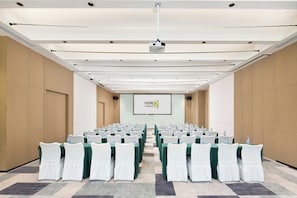 A conference room with rows of tables and chairs, a projector screen, and a large screen displaying 'HOME 2' on the wall.
