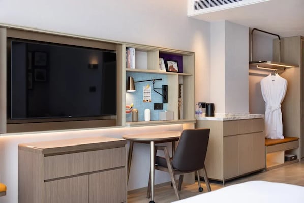 A modern hotel room with a large window, a desk with a chair, a small kitchenette, and a robe hanging on a hook.