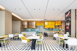 Interior de un restaurante moderno con suelo a cuadros, sillas amarillas y blancas, y una zona de cocina central.