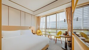 Ett hotellrum med en stor säng, ett skrivbord med en stol, ett litet bord med en lampa och utsikt över staden genom stora fönster.