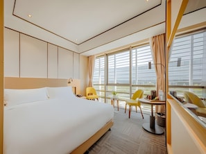 Habitación de hotel con una cama grande, un escritorio con silla, una mesita de noche con lámpara y vistas a la ciudad a través de amplios ventanales.