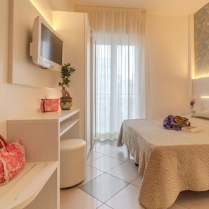 Una habitación de hotel con cama, televisor montado en la pared, una mesita con una planta y una ventana con cortinas.
