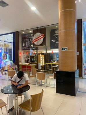 Un comedor moderno con un café de fondo, que presenta un pilar grande, disposición de mesas y una señal de salida visible.