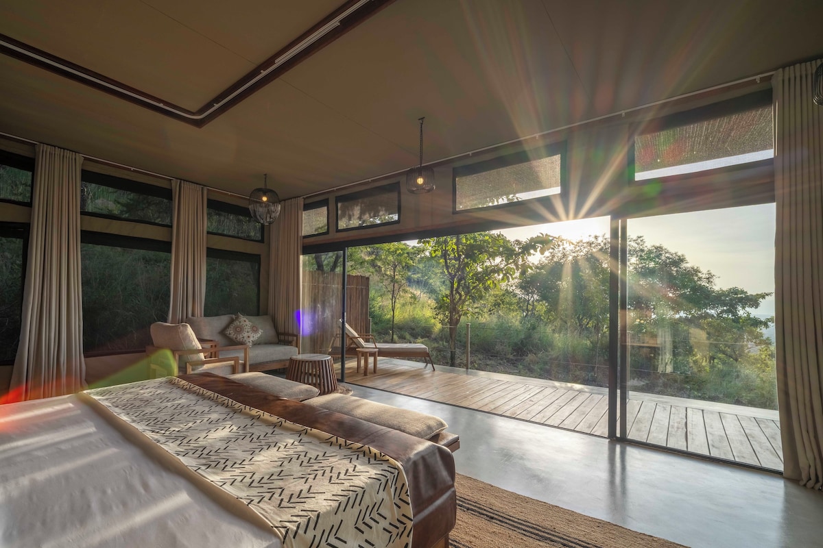 Quarto moderno com cama grande, sofá e varanda com vista para árvores.