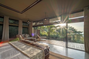 Quarto moderno com cama grande, sofá e varanda com vista para árvores.