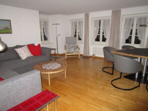 Un salon avec un canapé gris, un coussin rouge, une petite table ronde, un fauteuil gris et une table à manger avec des chaises.