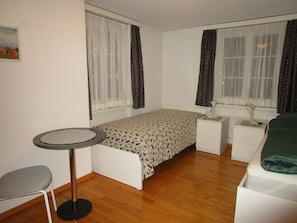 Una habitación pequeña y ordenada con una cama individual, un escritorio, una silla y dos ventanas con cortinas.