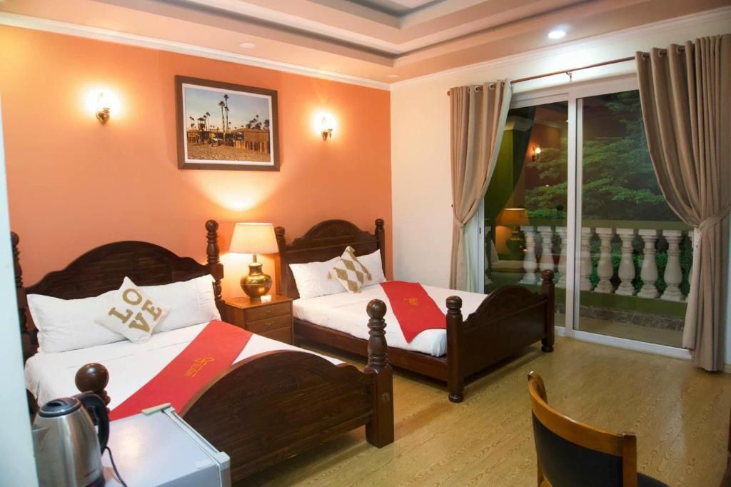 Habitación de hotel con dos camas de madera, una mesita de noche con lámpara, una silla y un balcón con cortinas.