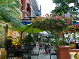 Un café al aire libre con mesas y sillas, rodeado de follaje y flores.