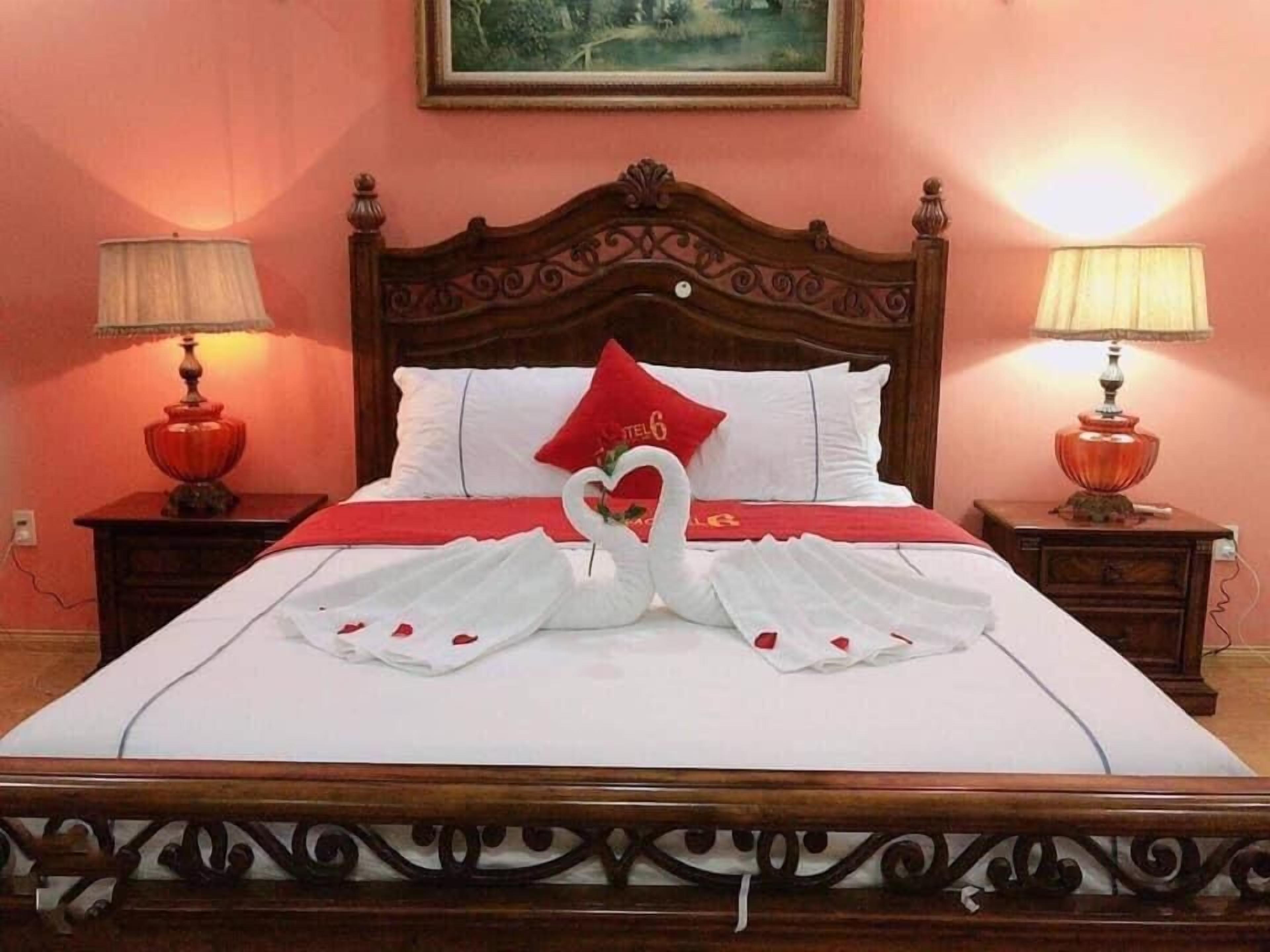 Cama com cabeceira decorativa, duas luminárias de cabeceira e um travesseiro vermelho com um detalhe em formato de coração.