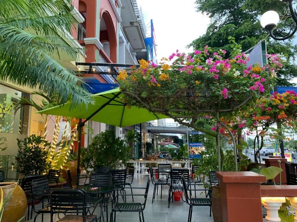 Un espacio de estar al aire libre con mesas y sillas, rodeado de follaje y plantas con flores.