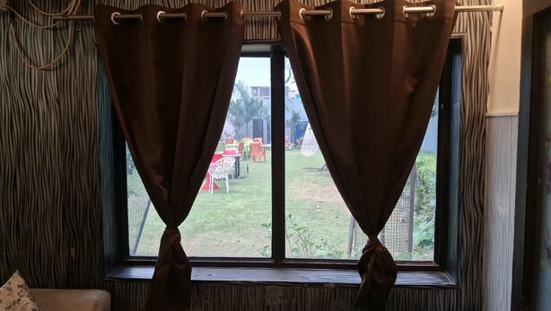 Una habitación con ventana, cortinas y vistas a un espacio exterior con mesas y sillas.