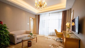 A hotel room with a sofa, a glass coffee table, a desk with a TV, a potted plant, and a large window with curtains. - Vienna International Hotel (Foshan)