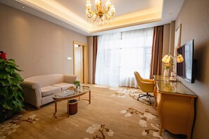 Ein Hotelzimmer mit einer Couch, einem Glastisch, einem Schreibtisch mit Fernseher, einer Topfpflanze und einem großen Fenster mit Vorhängen.