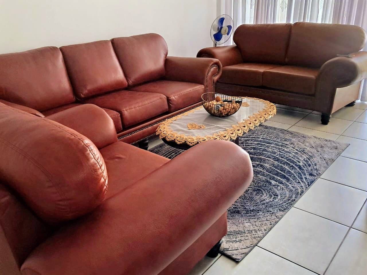 En brun lædersofa med et vævet sofabord, en ventilator og et mønstret tæppe i et rum med hvide vægge og flisegulv.