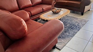 Un canapé en cuir marron, une table basse en osier, un ventilateur et un tapis à motifs dans une pièce aux murs blancs et au sol carrelé.