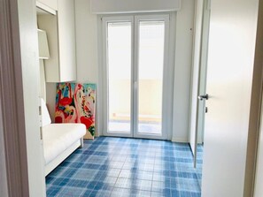 Een kamer met een wit bed, een kleurrijk schilderij en een blauw geruit vloertje.