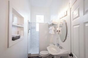 Casa de banho compacta com lavatório branco, sanita e zona de duche. Há uma janela a permitir luz natural e um porta-toalhas com toalhas dobradas.