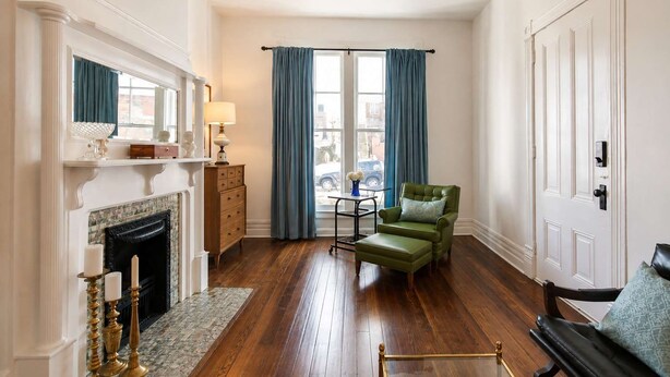 A room with a fireplace, a green armchair, a small table with a vase, and a view through a window with blue curtains. - Relaxing Apt near Cherokee Park and Frankfort Ave! (Louisville)