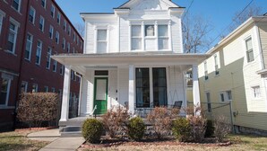 A two-story white house with a green door, a front porch with chairs, and a small garden. - Relaxing Apt near Cherokee Park and Frankfort Ave! (Louisville)