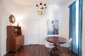 Uma sala de jantar com uma mesa redonda, cadeiras brancas, um armário de madeira, um lustre e um quadro na parede.