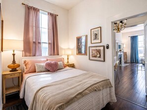 Quarto com cama, abajures, janela com cortinas e quadros nas paredes.