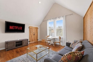 A modern living room with a flat-screen TV, a glass coffee table, a gray sofa, and a dining area with a table and chairs. - Cozy Phoenix Hill suite in the heart of Louisville (Louisville)