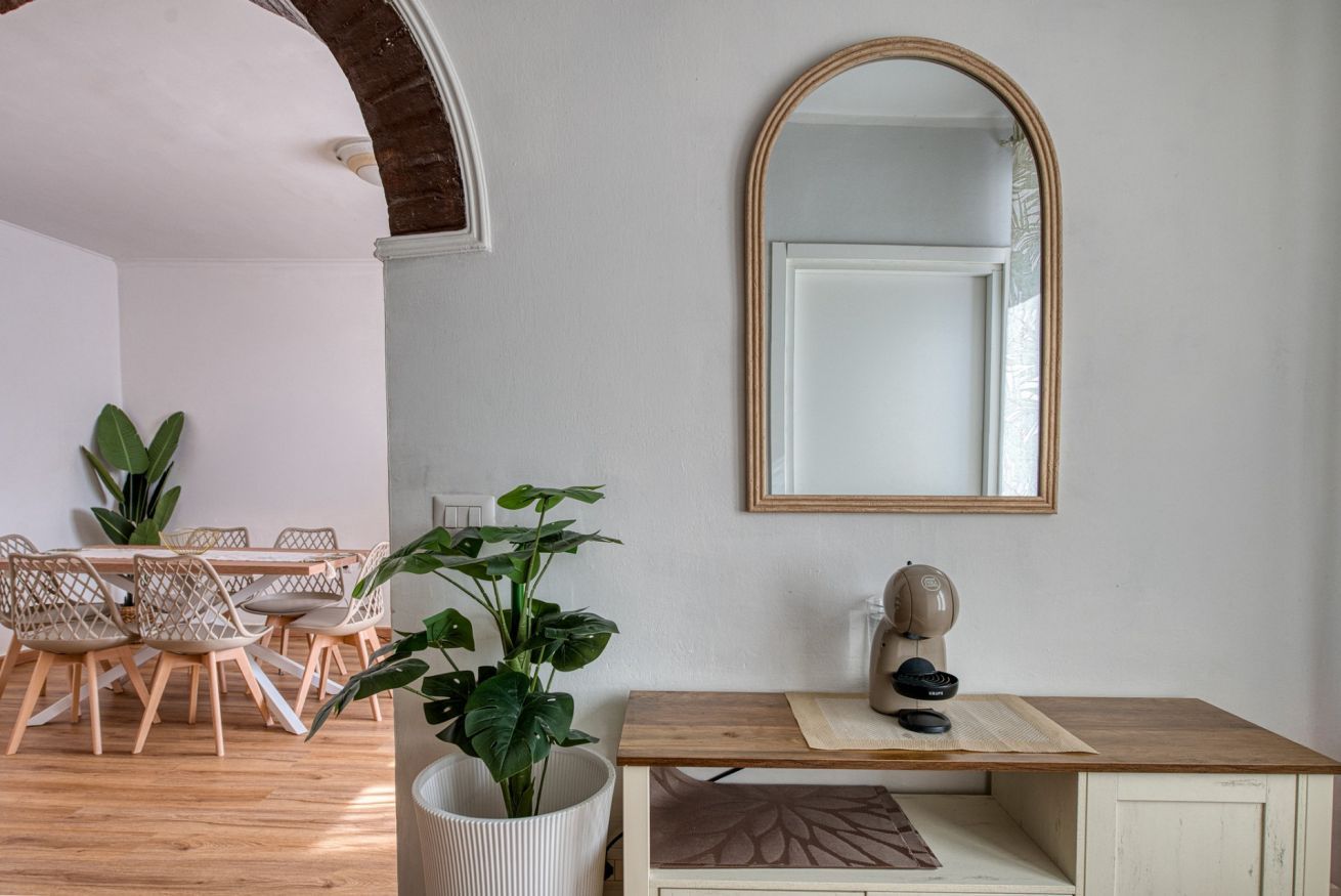 A room with a dining table, chairs, a potted plant, a desk with a mirror, and a wall-mounted lamp.